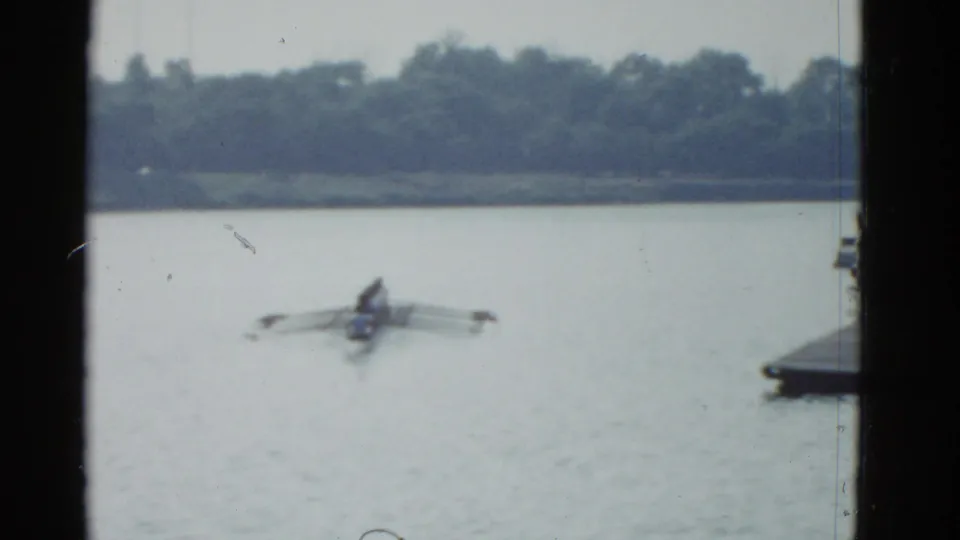 1975 Boston-area 8mm frame showing open lake water with a small rowing craft and oars, with a distant wooded shoreline behind it.