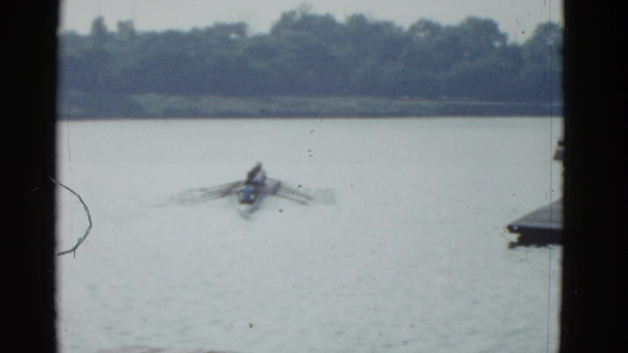 Distant rower in a shell moving across a lake with trees beyond and dark frame edges.