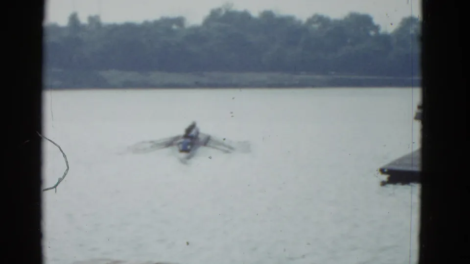 1975 Boston-area 8mm frame showing a distant rower on a lake, with oars spread wide, a tree-lined shore, and dark edges in the foreground.