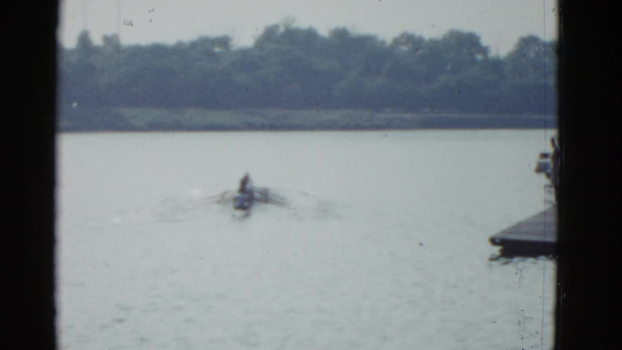 1975 Boston-area 8mm frame with a small rowing shell centered on the lake, a tree line in the distance, and a dock edge at right.