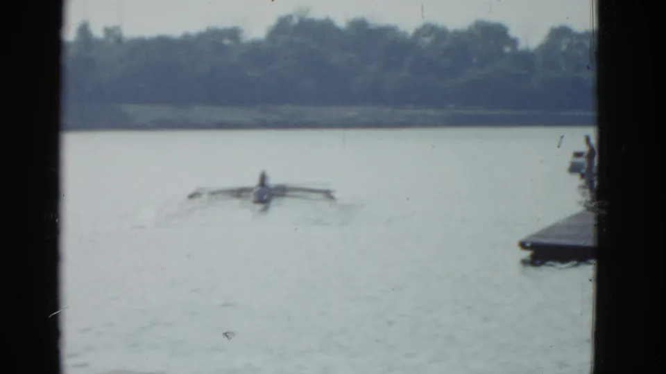 1975 Boston-area 8mm frame showing the rower farther across the lake, with soft focus, pale water, and a wooded shoreline behind.