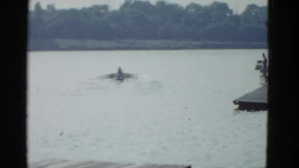 1975 Boston-area 8mm frame of a distant sculler on calm water, with broad oars, light wake, shoreline trees, and dark film-frame edges.