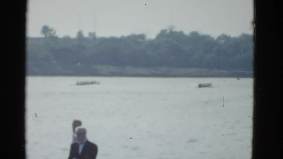 A distant rowing shell crosses the Boston waterfront while two shoreline observers stand in the lower foreground of the 1975 home movie.