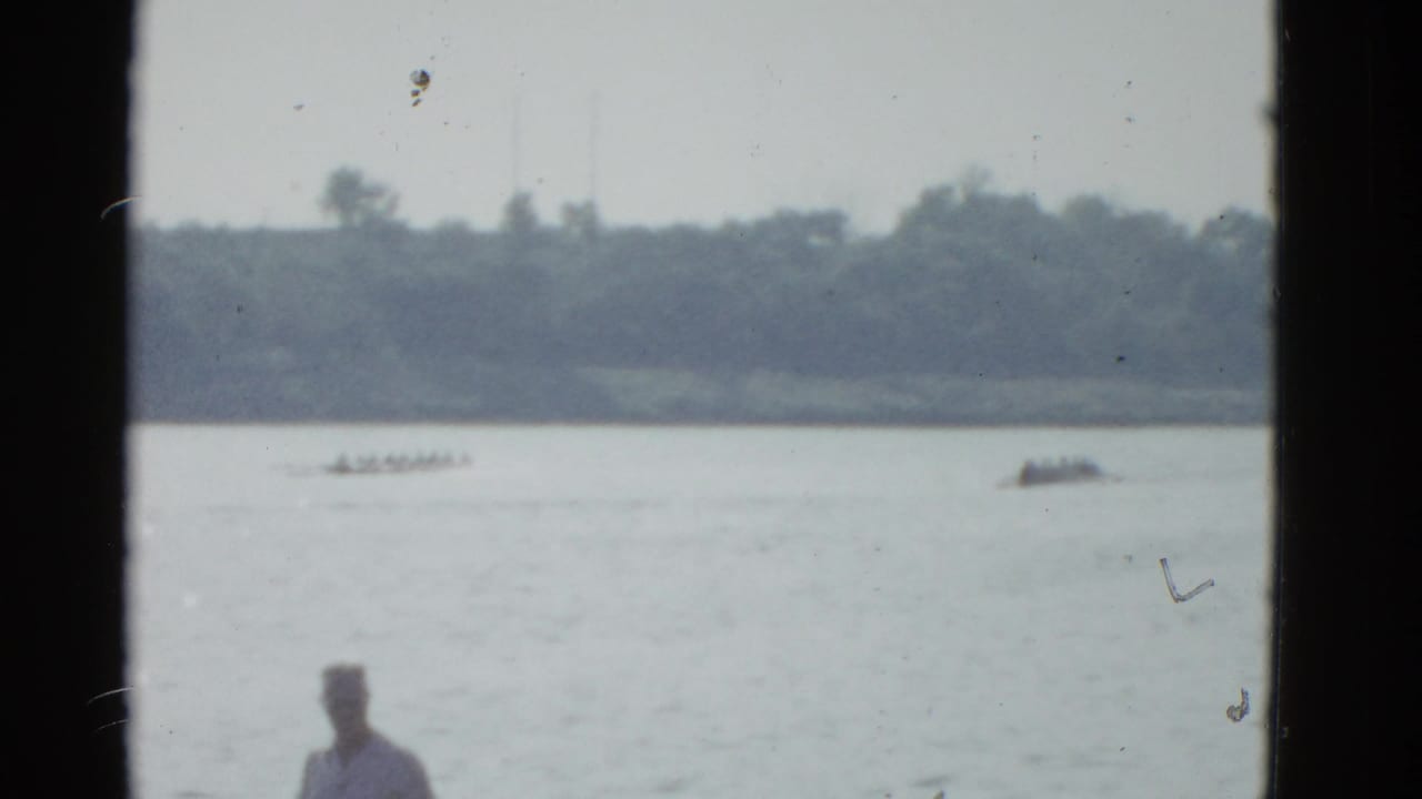 1975 Boston-area 8mm frame showing distant rowing shells on calm water with a shoreline spectator partly visible in front.