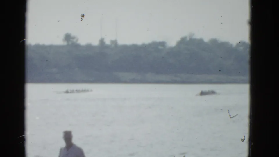 1975 Boston-area 8mm frame showing distant rowing shells on calm water with a shoreline spectator partly visible in front.