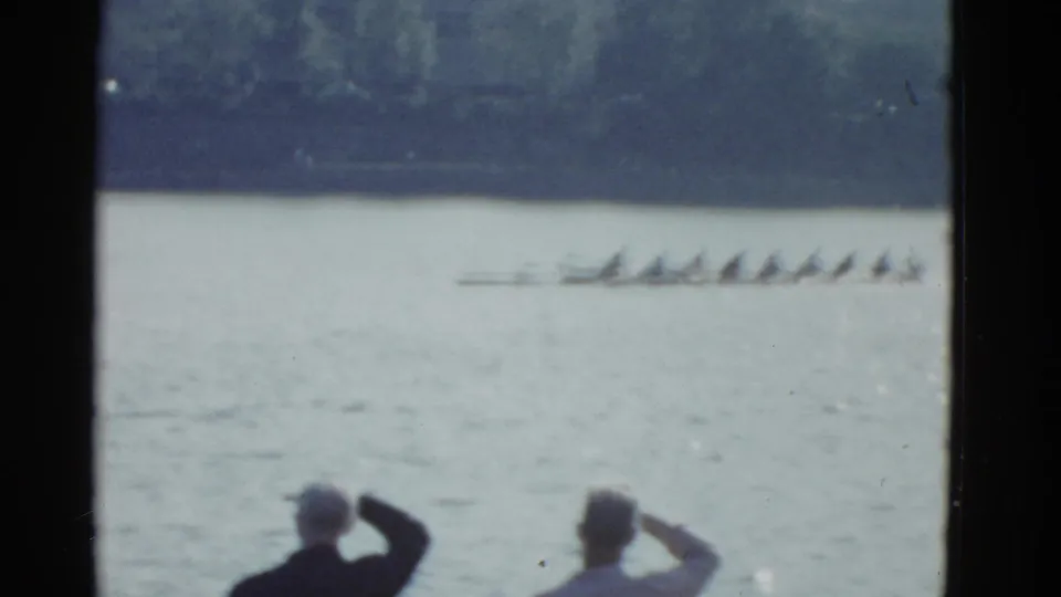 Two shoreline spectators watch a long racing shell on the river in metadata-supported 1975 Boston home-movie footage.