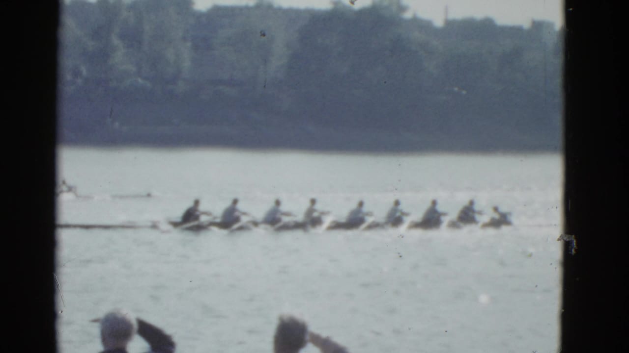 Two rowing shells move across calm water with spectators partly visible at the bottom edge.