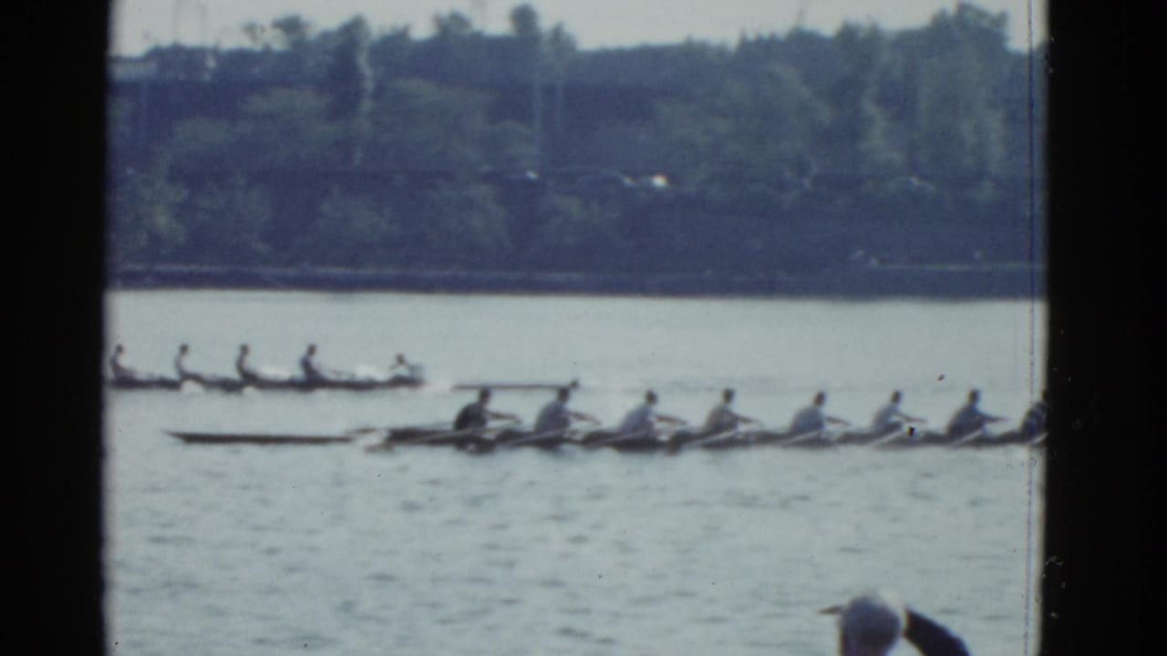 1975 Boston 8mm frame with multiple rowing shells on the water, oars extended as teams pass near a tree-lined shore.