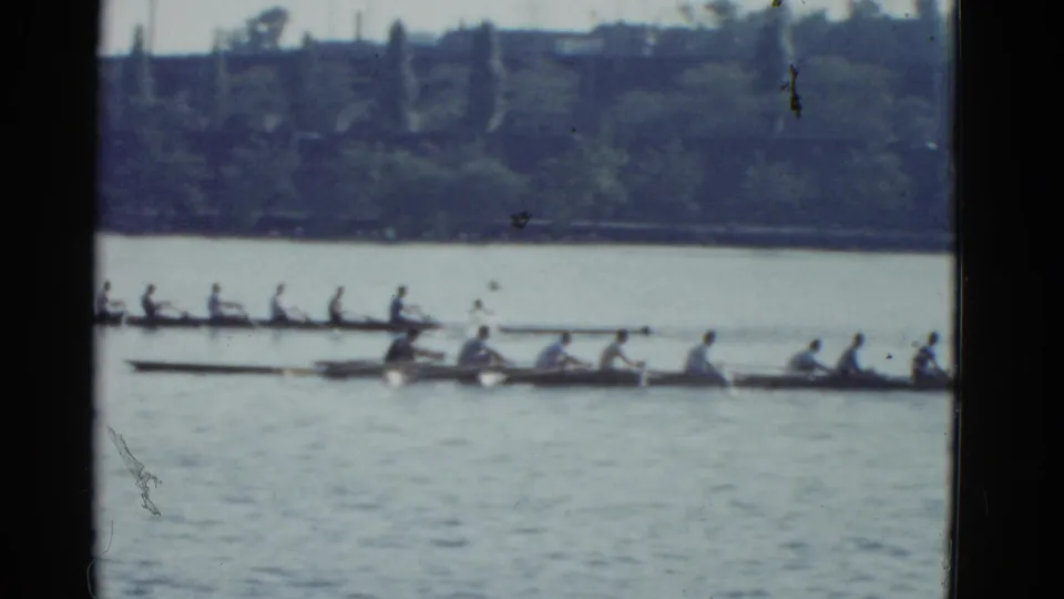 1975 Boston 8mm frame showing two crew shells crossing the water, their rowers seated in line against a distant shoreline.