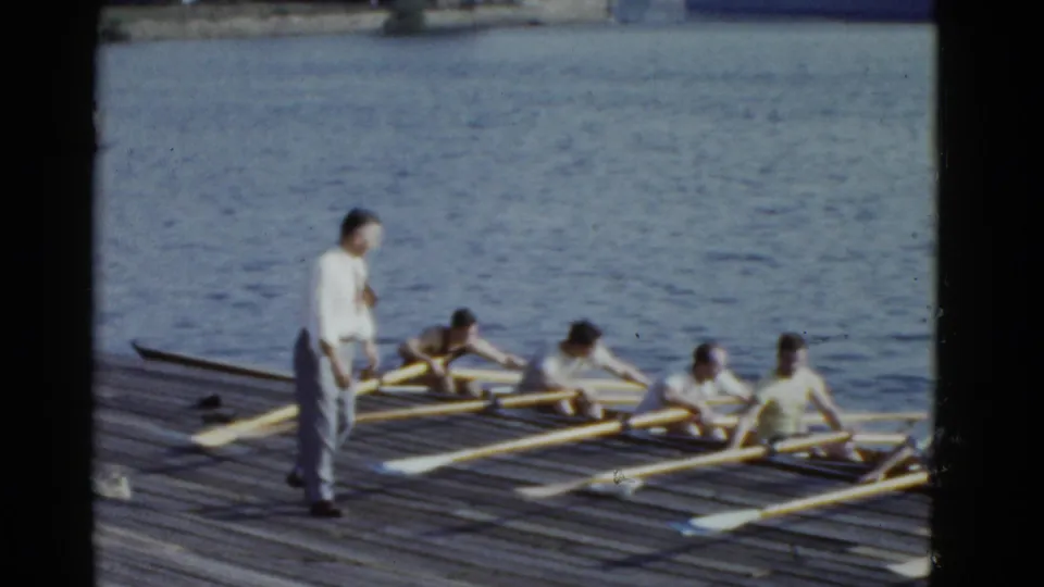 1975 Boston 8mm frame showing rowers beside a wooden dock, with an adult standing near the shell and oars laid across the water.