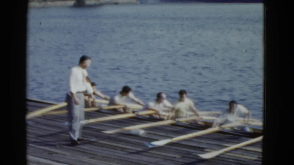 1975 Boston home-movie frame showing men in a rowing shell near a wooden dock, with a standing observer beside calm water.