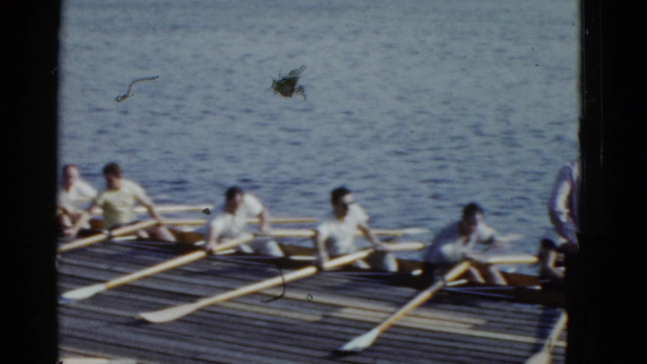 Men in light shirts sit in a long rowing shell at a Boston waterfront dock, holding wooden oars during a 1975 practice moment.