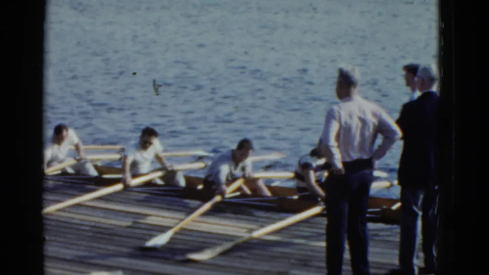 Boston 1975 rowing practice continues as men stand on the dock and rowers adjust in the shell with oars spread across the waterline.