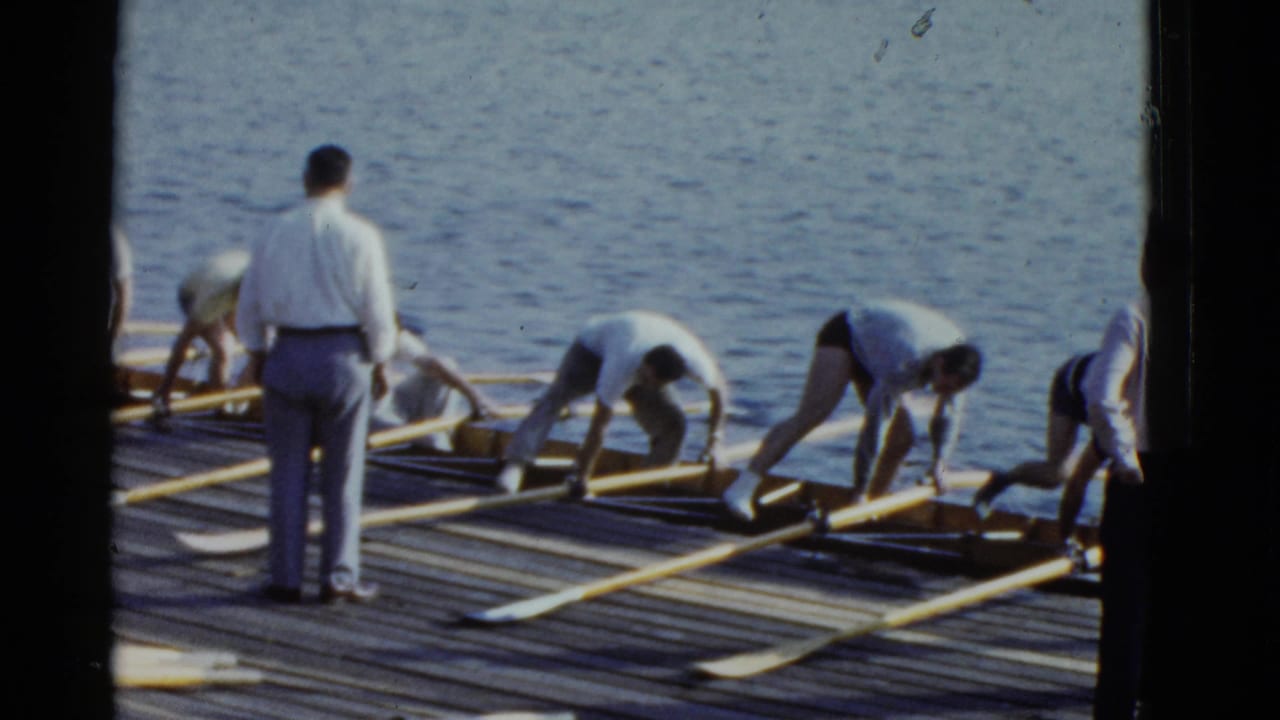 Rowers prepare long oars beside a wooden dock while a man in a white shirt stands nearby.