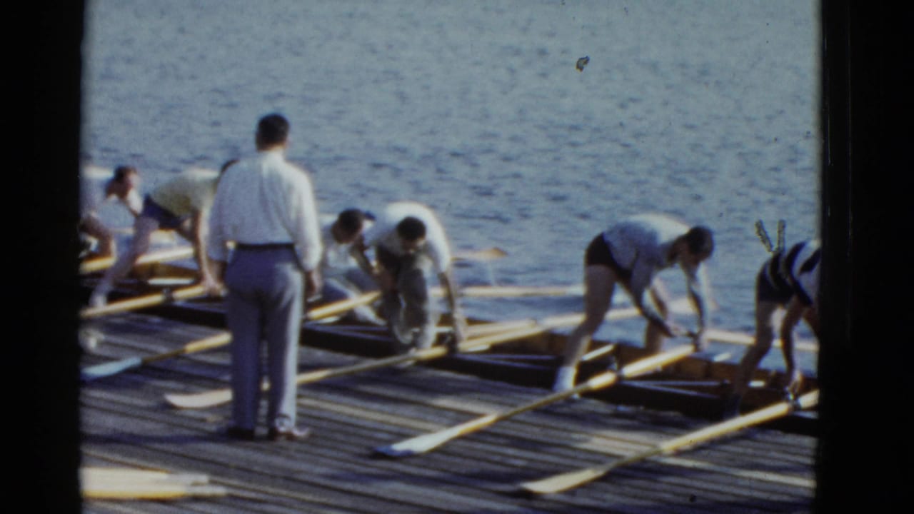 Adults on a Boston dock in 1975 appear to prepare a rowing shell, with long oars laid out and water filling the background.
