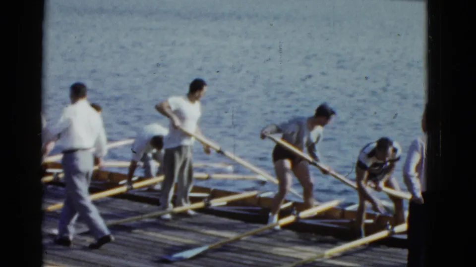 1975 8mm home-movie frame showing Boston rowers repositioning long oars on a wooden dock with calm water behind them.