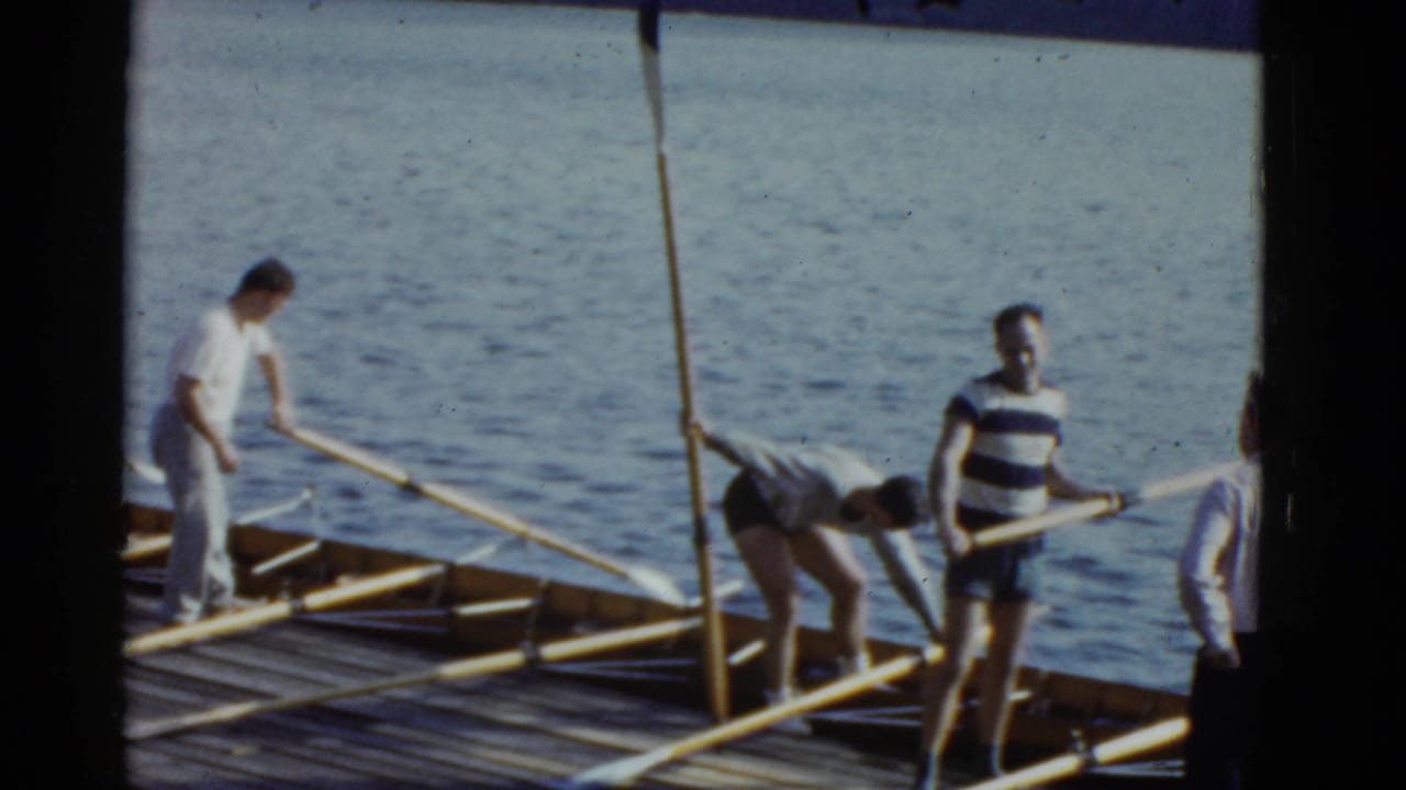 Men in athletic clothes prepare rowing oars on a wooden dock in 1975 Boston-area home-movie footage.