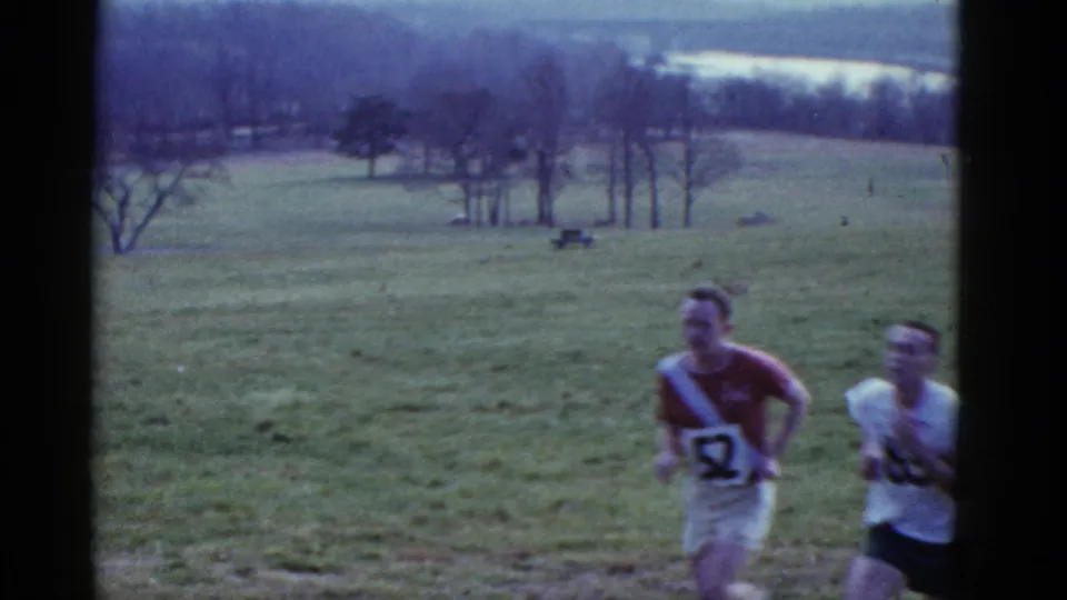1975 Boston-area 8mm frame with two runners wearing numbered race tops crossing an open grassy field.