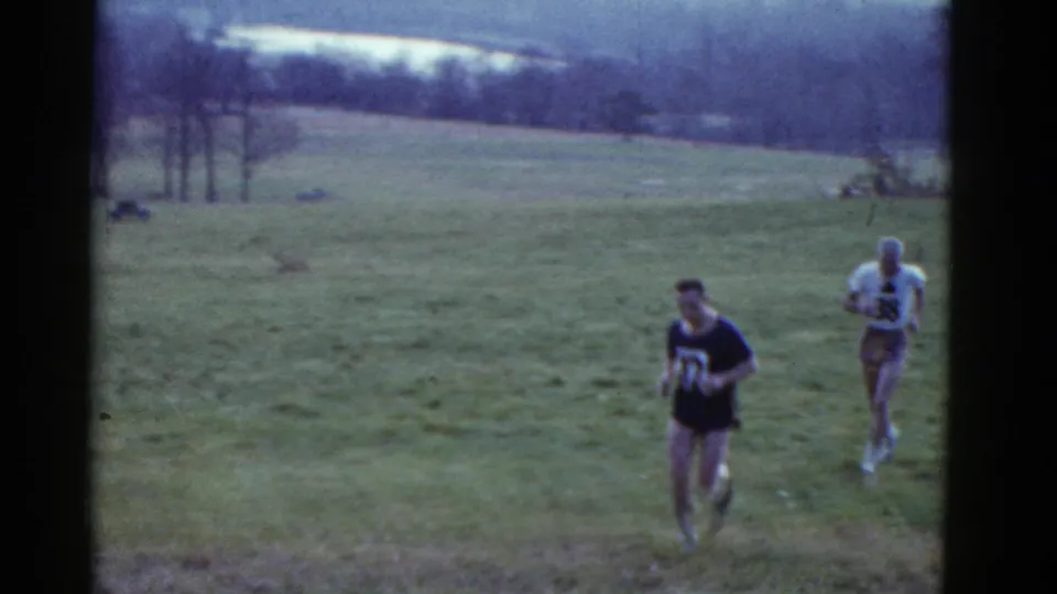 In 1975 Boston home-movie footage, two adult male runners wearing race numbers cross an open grassy field.