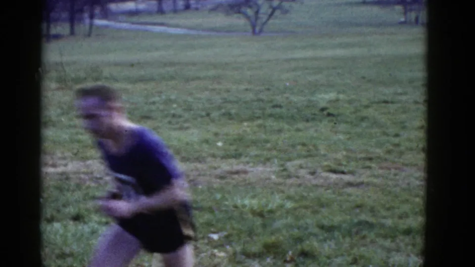 A male runner in a dark numbered shirt moves close past the handheld 8mm camera, with the grassy field behind him.