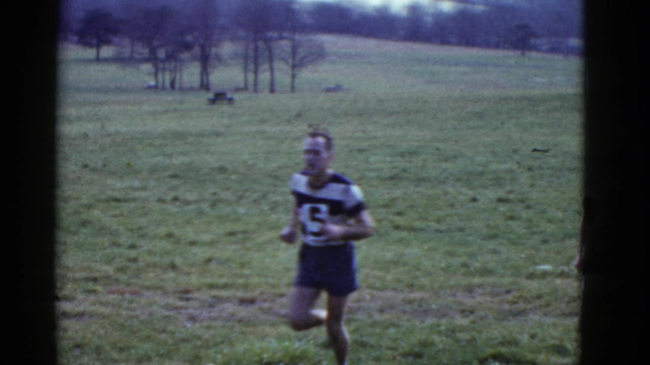 A male runner in a dark uniform and numbered bib passes through the Boston-area grassy race course.