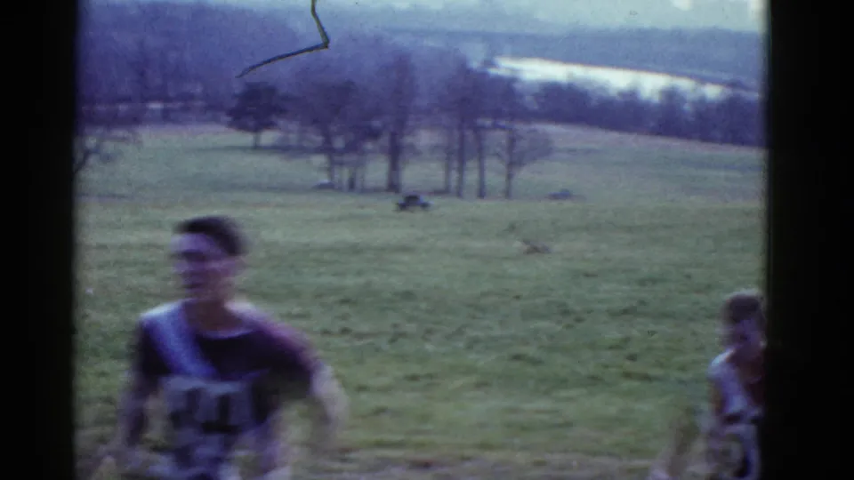 1975 Boston-area 8mm frame showing a blurred male runner in a numbered athletic jersey moving across an open grassy field.
