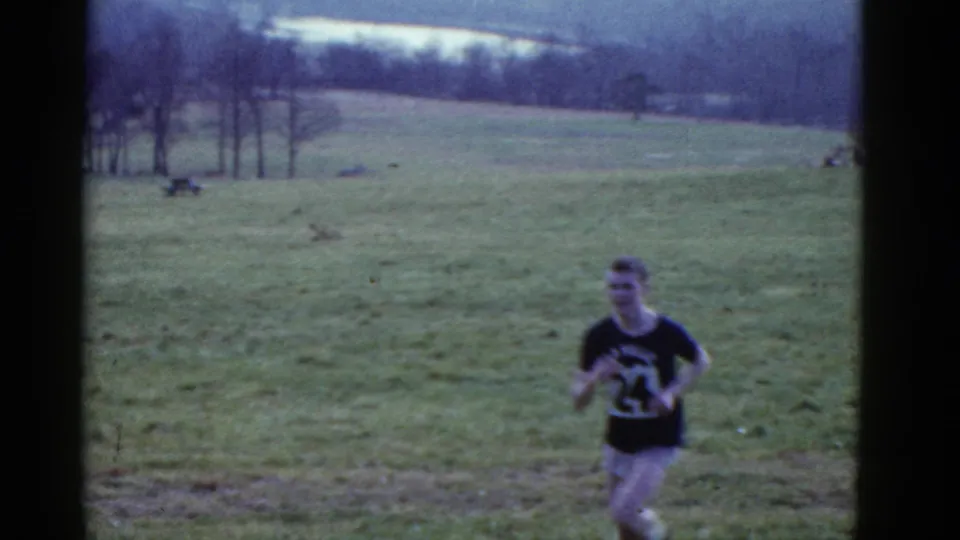 1975 Boston-area home-movie frame of a male runner in a dark numbered shirt approaching across a sloped grassy field.
