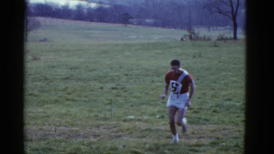 1975 Boston-area 8mm frame showing a male runner in a maroon and white number 53 jersey crossing a grassy field.