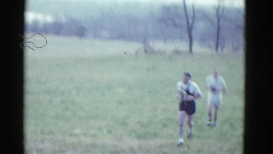 1975 8mm home-movie frame of two runners crossing a grass field in Boston, with bare trees and soft distant hills behind them.