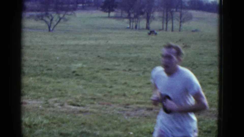A runner in a white shirt moves past the camera in a 1975 Boston cross-country scene, with open grass and bare trees in the background.