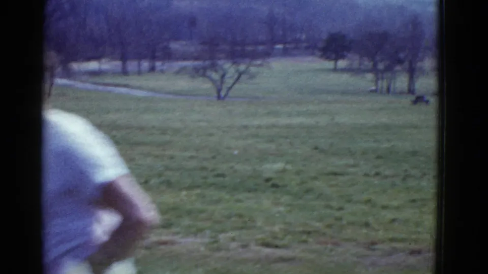 1975 8mm frame from the Boston-listed clip showing a partial runner passing the camera near an open field with bare trees and distant woods.