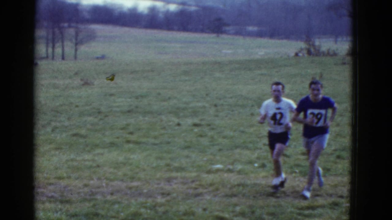 Two adult runners, one in a white shirt numbered 42 and one in a blue shirt numbered 39, move across a grassy field in the 1975 Boston-listed clip.