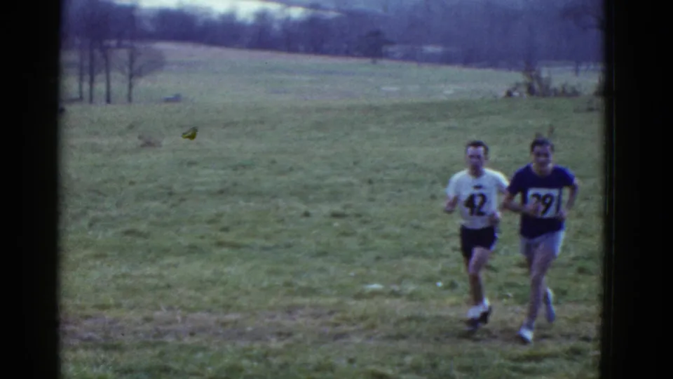 Two adult runners, one in a white shirt numbered 42 and one in a blue shirt numbered 39, move across a grassy field in the 1975 Boston-listed clip.