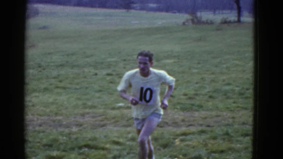 A runner in a yellow shirt with number 10 moves across the grassy course, with open field and distant trees behind him in the 1975 Boston-listed footage.