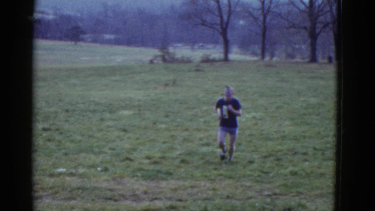 A runner in a dark numbered shirt approaches from the distance across an open grassy field near Boston in 1975 footage.