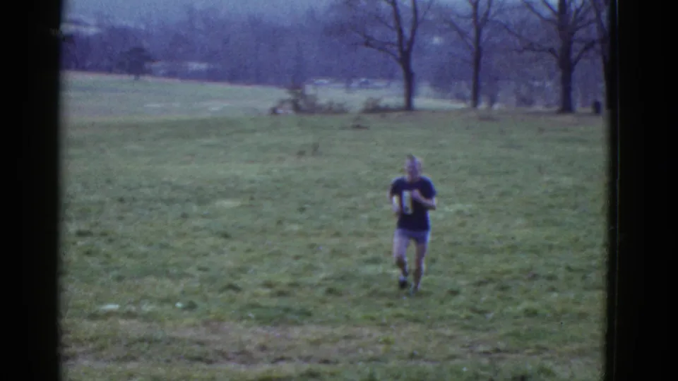 A runner in a dark numbered shirt approaches from the distance across an open grassy field near Boston in 1975 footage.