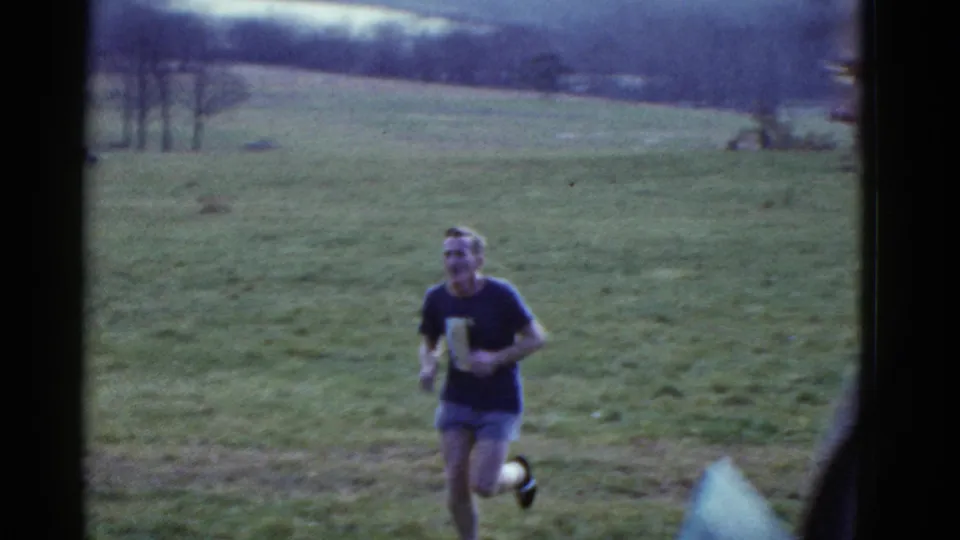 A dark-shirted runner passes closer in the grassy field as a spectator or bystander appears at the right edge of the 8mm frame.
