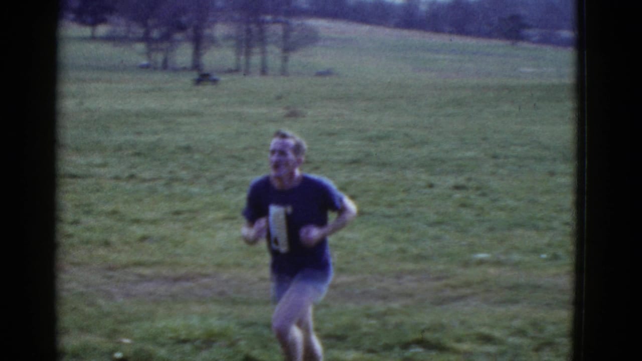 Man running across a grassy park with open field and trees behind him.