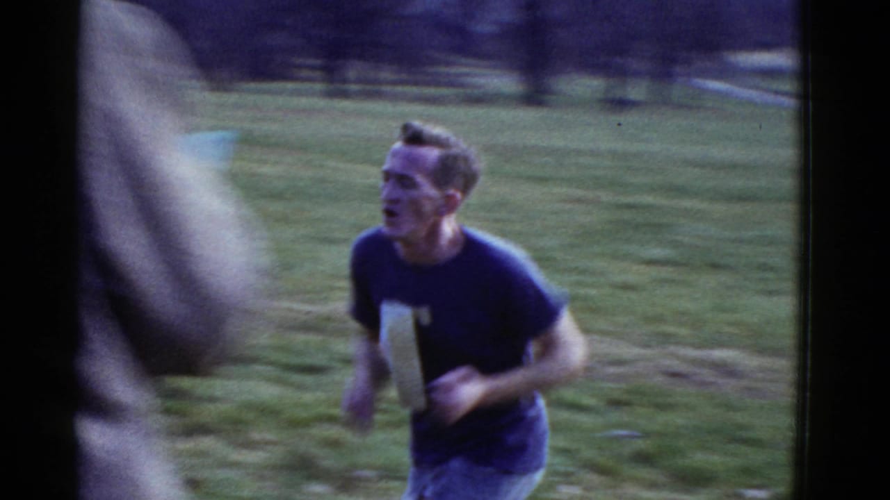 Boston-area 1975 home-movie frame showing a runner on grass with a blurred person crossing the foreground.