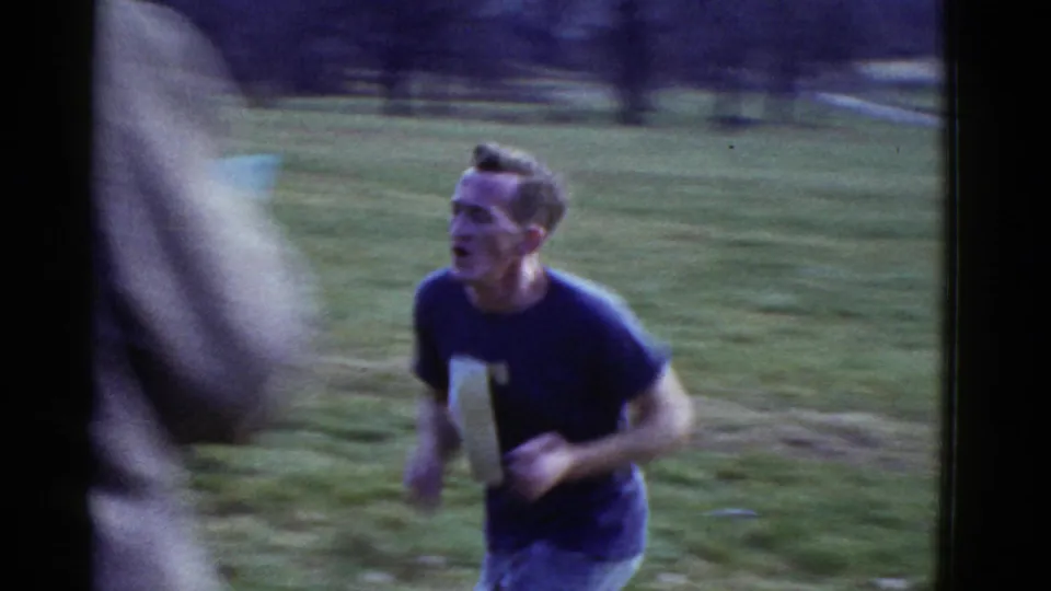 Boston-area 1975 home-movie frame showing a runner on grass with a blurred person crossing the foreground.