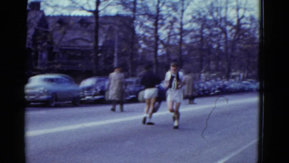 1975 Boston-area road-race view with runners moving down a tree-lined street past parked cars and spectators.