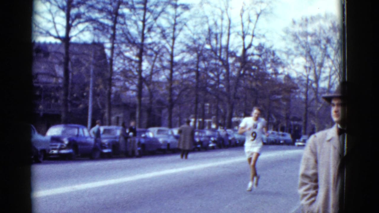 1975 Boston road-race scene with a runner in a white number 9 jersey moving toward camera near spectators, parked cars, and bare trees.