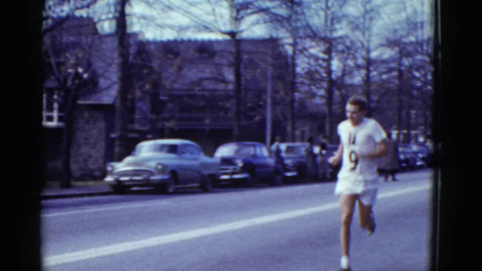 Boston 1975 8mm frame showing a male runner in a white number 9 jersey passing a curb lined with mid-century cars and bare trees.