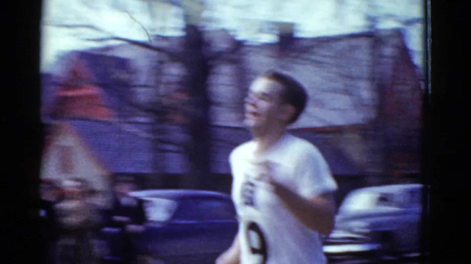 Close handheld 8mm view of a 1975 Boston runner in a white number 9 jersey, with motion blur, parked cars, and buildings behind him.