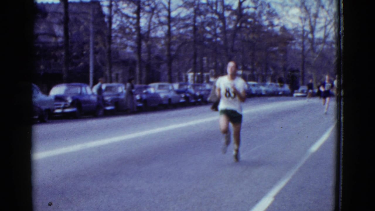 Runner in a numbered shirt approaches on a city street lined with parked cars and bare trees.