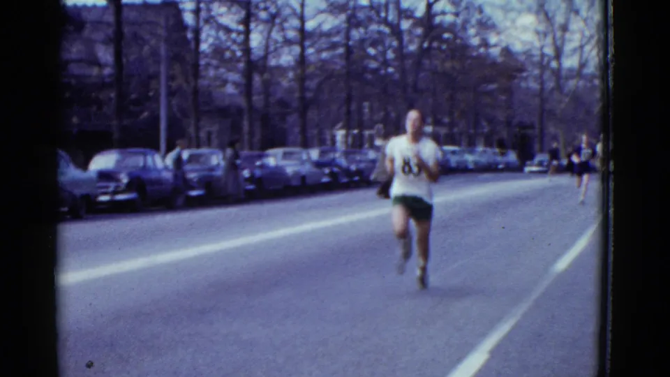1975 Boston 8mm frame of a marathon runner approaching along a broad street with parked cars, bare trees, and spectators in the distance.