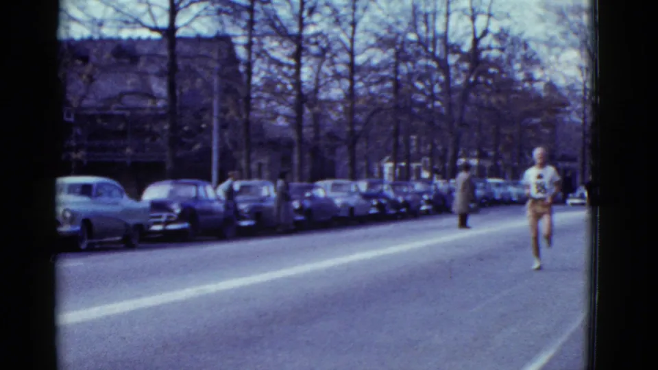 1975 Boston 8mm frame showing a marathon runner farther down the street, with a pedestrian, parked cars, and bare trees along the route.