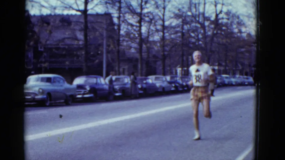 1975 Boston 8mm frame of a runner with bib number 88 approaching on a city street lined with parked cars and bare trees.