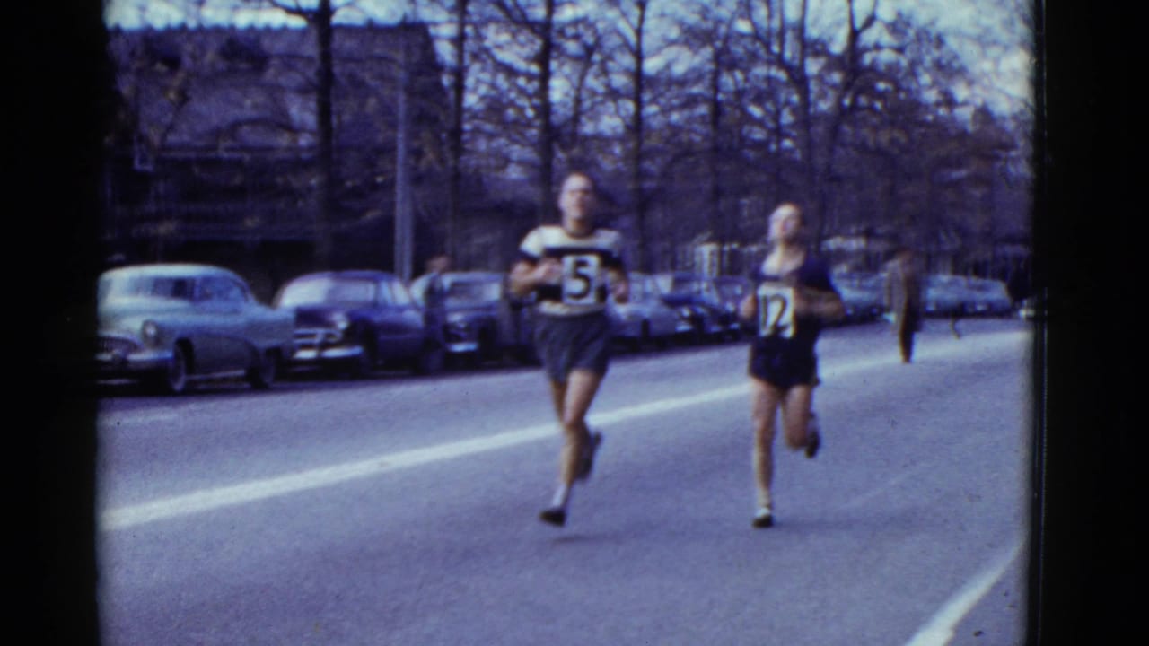 Two male runners, one wearing bib 5 and another wearing bib 12, pace along a Boston street in 1975 archival home-movie footage.