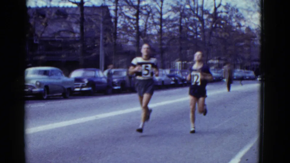 Two male runners, one wearing bib 5 and another wearing bib 12, pace along a Boston street in 1975 archival home-movie footage.